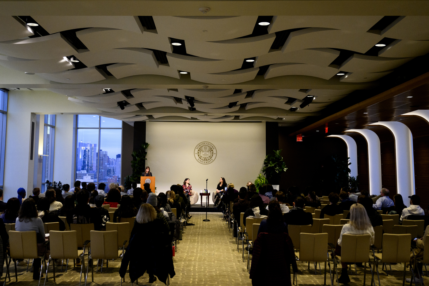 Audience members sit in an auditorium, listening to people talking on a stage.