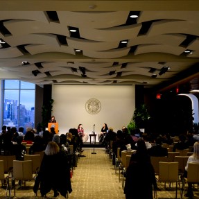 Audience members sit in an auditorium, listening to people talking on a stage.