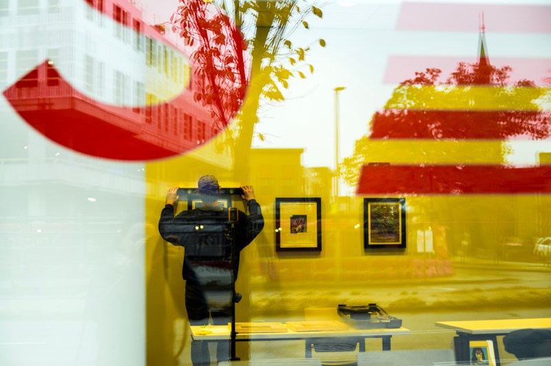 Person hanging art on the wall of the exhibit, captured through a glass window. 