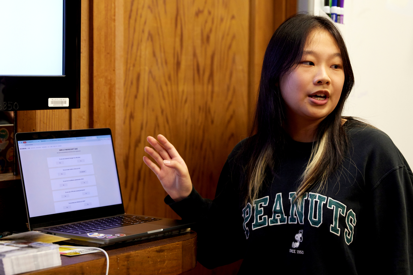 Student wearing a black sweater that says 'Peanuts' on it in green letters gesturing  to their open laptop. 