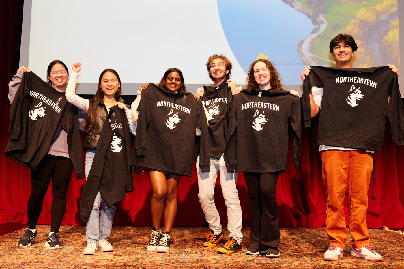 A group of six students holding Northeastern branded long-sleeve shirts up and smiling. 