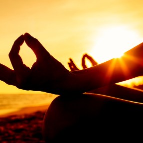 Silhouette of a person at sunset sitting cross legged with their hands resting in their lap, thumb and index fingers pressed together and palms upwards in Gyan mudra.