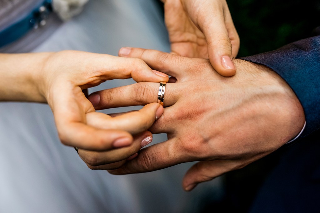 Bride slipping a wedding ring on the ring finger of a groom.