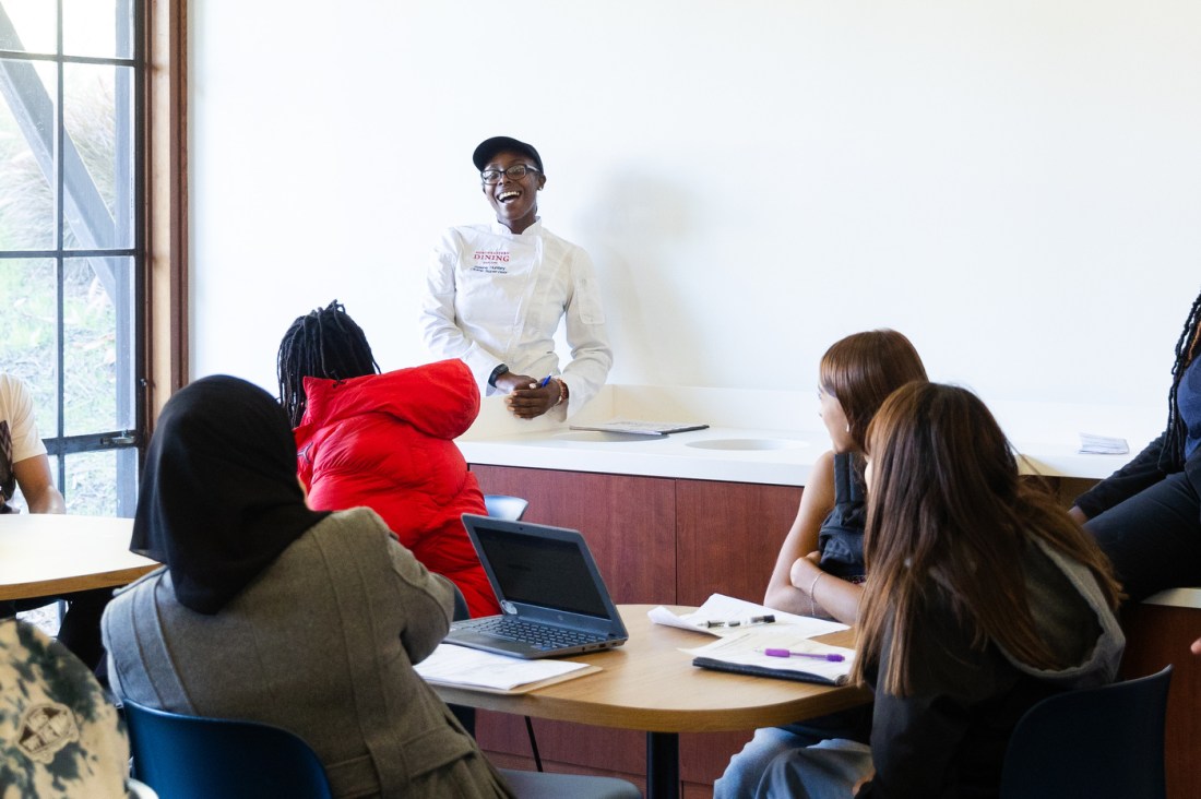 Northeastern Dining worker in their white uniform smiling while talking to students sitting around tables with papers and pencils. 