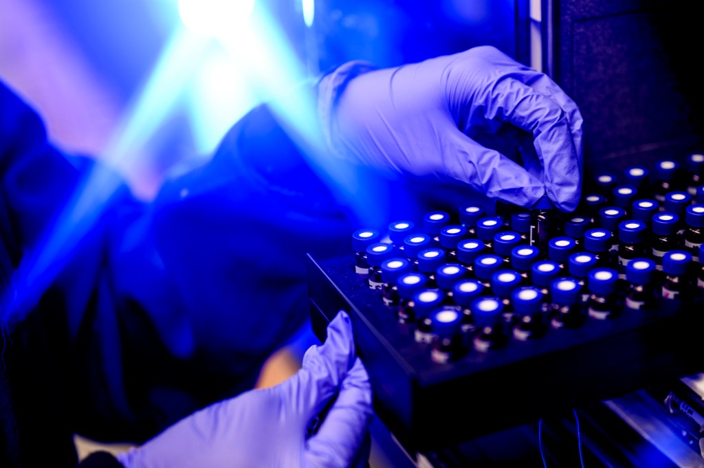 Hands wearing purple-colored gloves place a vial into a container in a blue-lit room.