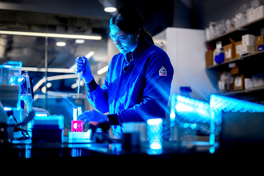 A person wearing a blue lab coat and goggles works with test tubes inside a laboratory.