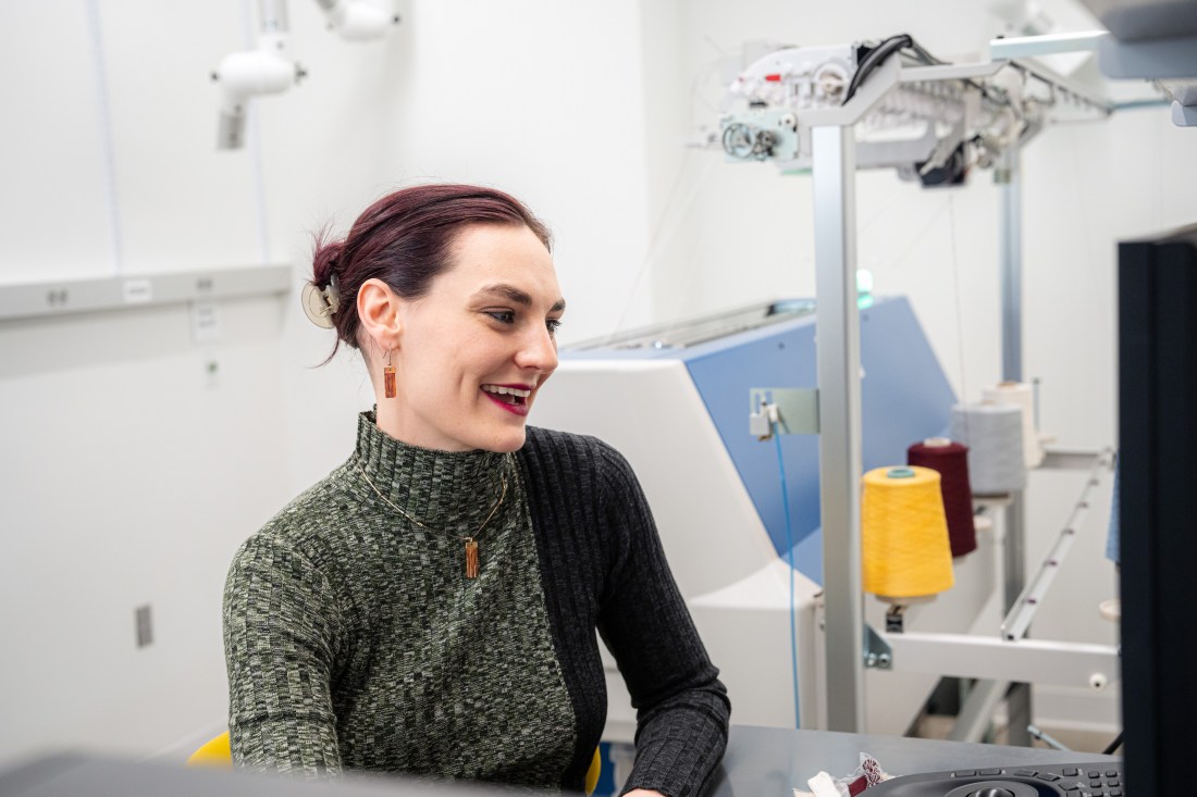 Professor Megan Hofmann sits in front of a large machine in a white room. Differently colored spools of yarn sit along the right side, attached to the machine.