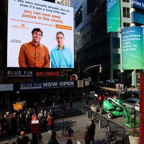 A PSA about the Adult Surivvors act displaying two people and some text playing on a screen in Times Square.