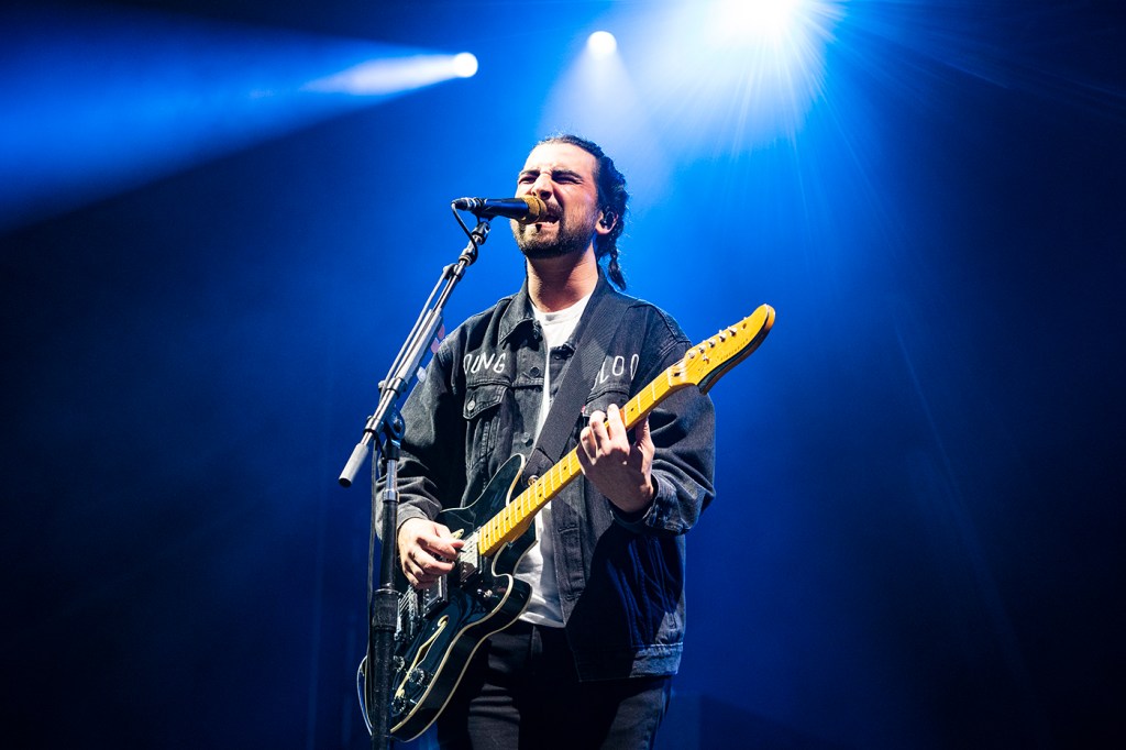 Noah Kahan playing guitar while singing into a microphone on stage under blue lights.