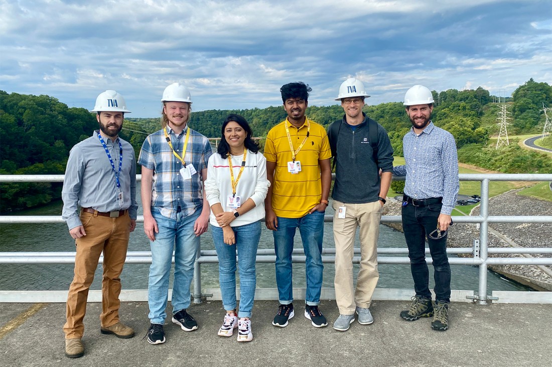 A group of six scientists and doctoral students on top of a dam in Tennessee, four wearing hard hats.