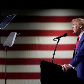 Donald Trump speaking at a microphone during a rally in front of a large United States flag.