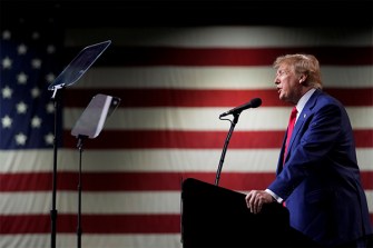 Donald Trump speaking at a microphone during a rally in front of a large United States flag.