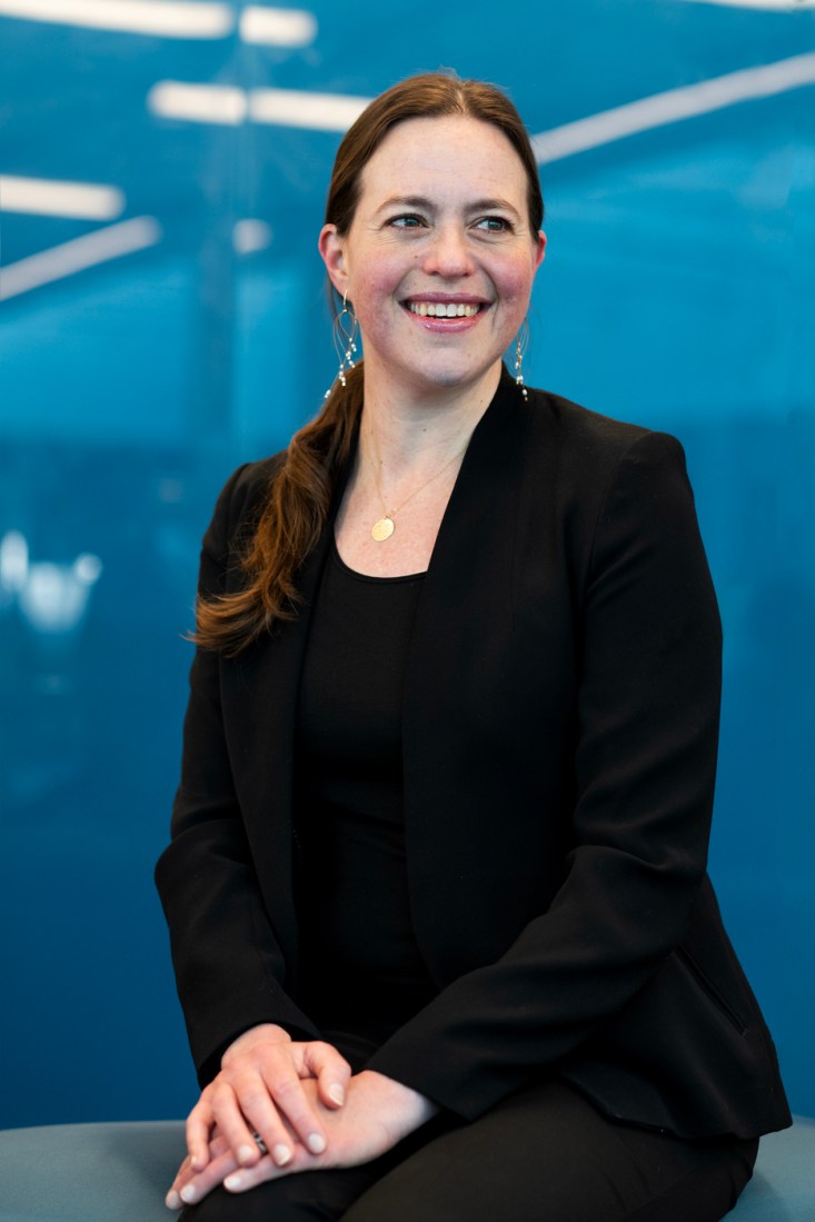 A woman in a black suit sits for a portrait against a blue background.