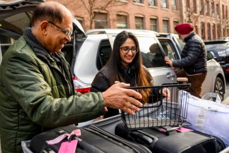A person wearing a green jacket helps another person load items into a large bin outside on a sunny day.