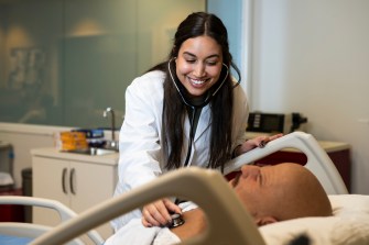 A person wearing a medical coat uses a stethoscope on a patient lying in a bed.