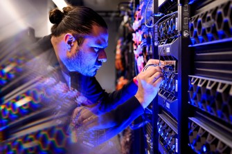 A person works in a blue-lit room wearing a buttoned shirt and handling multiple wires.
