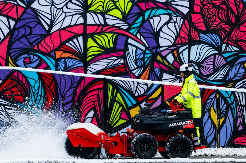 A person wearing a neon-yellow jacket uses a snowplow to clear a path in front of a multi-colored mural.