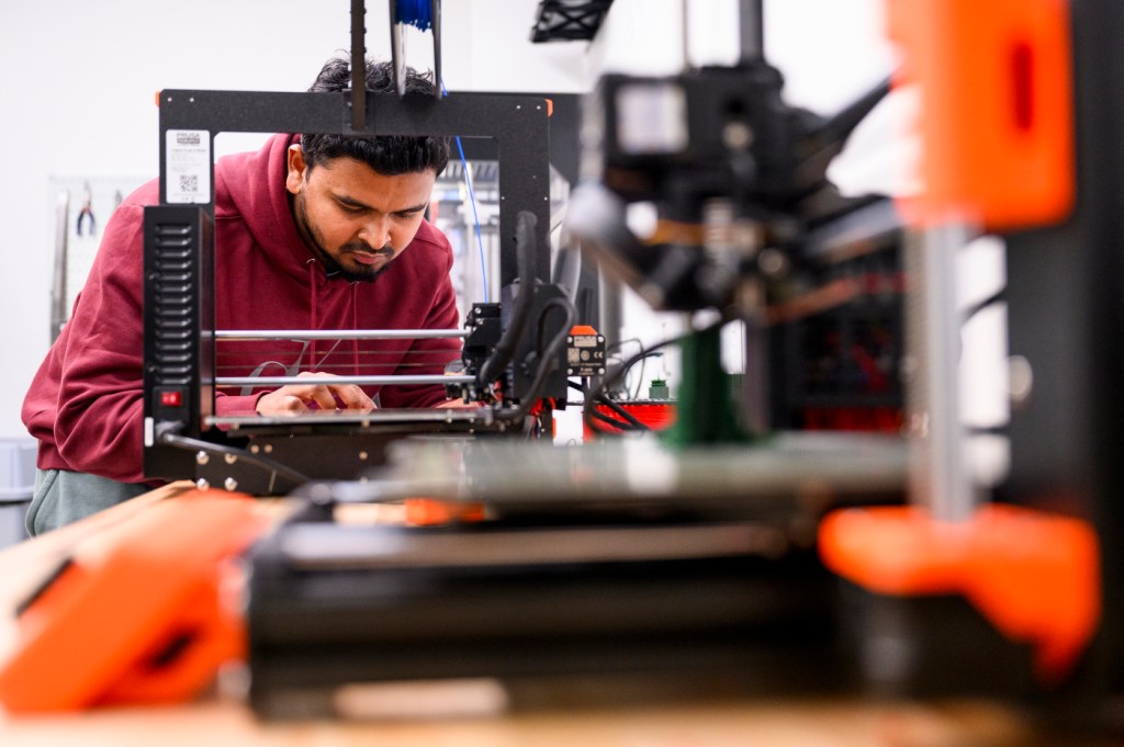 A person wearing a red hoodie works with a 3D printer.