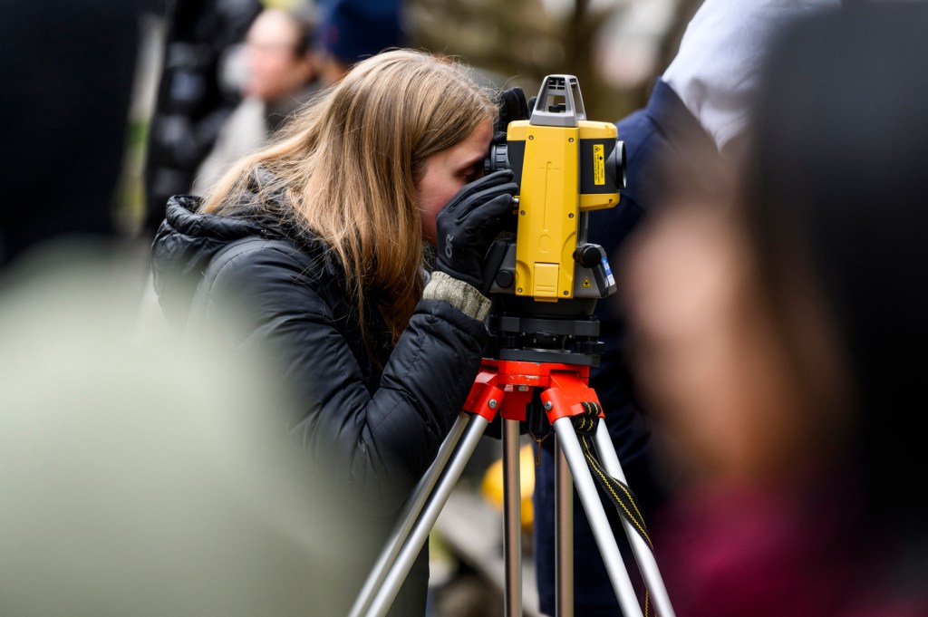 A person wearing a black jacket and gloves looks through a level (optical instrument) to conduct an elevation survey.