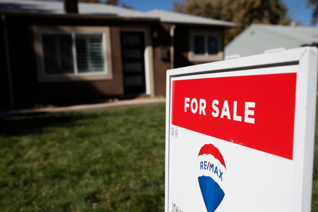 A Re/Max 'For Sale' sign on the lawn outside of a one-story brick house.