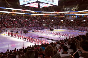 A brightly lit, large ice arena sold out with audience members watching a hockey game with players lined up on the ice inside the rink.