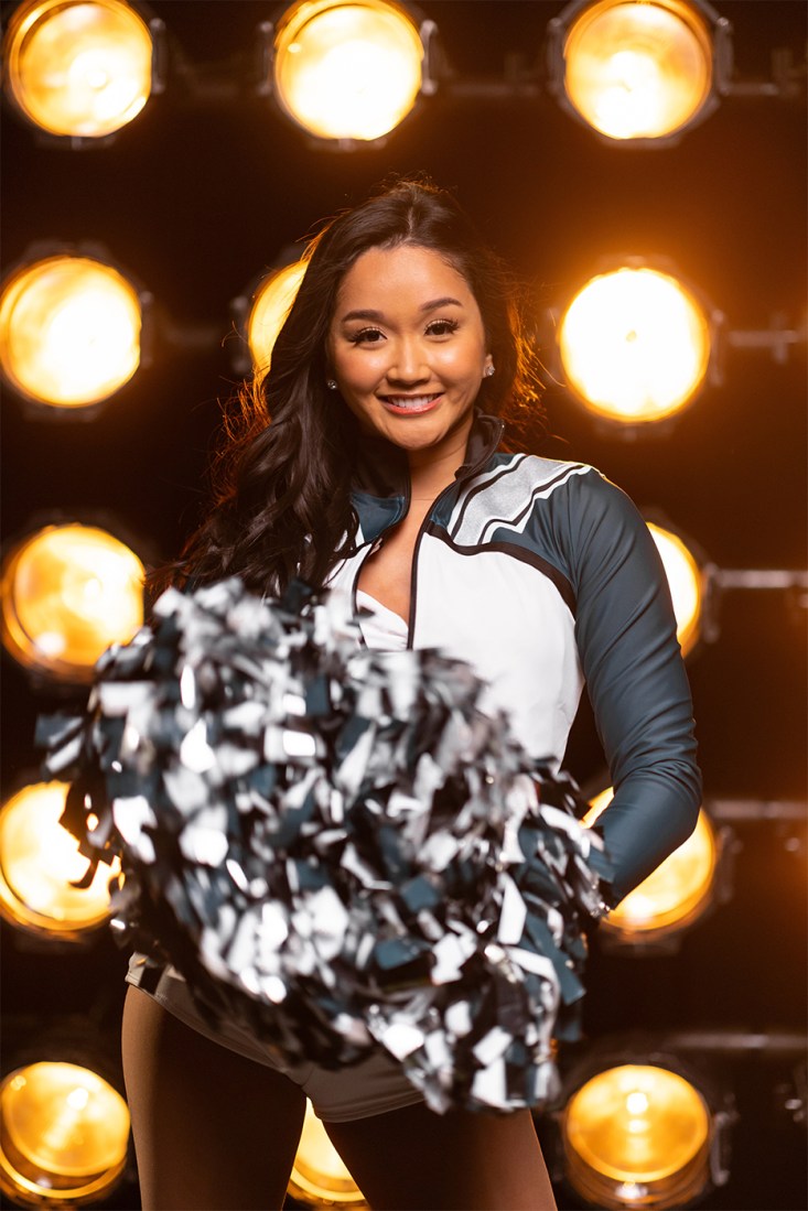 Chelsea Pe Benito in her Philadelphia Eagles cheerleader outfit holding pom poms in front of bright studio lights. 