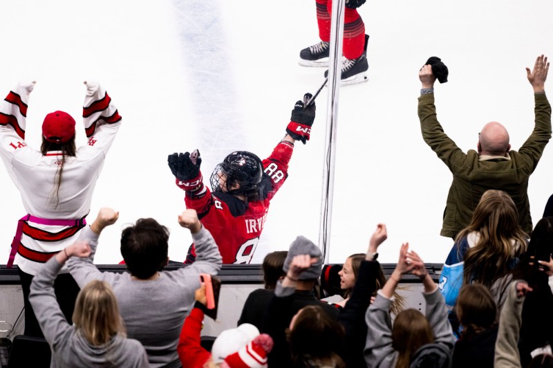 Fans cheering and banging on the side line glass while women's hockey players play on the ice.