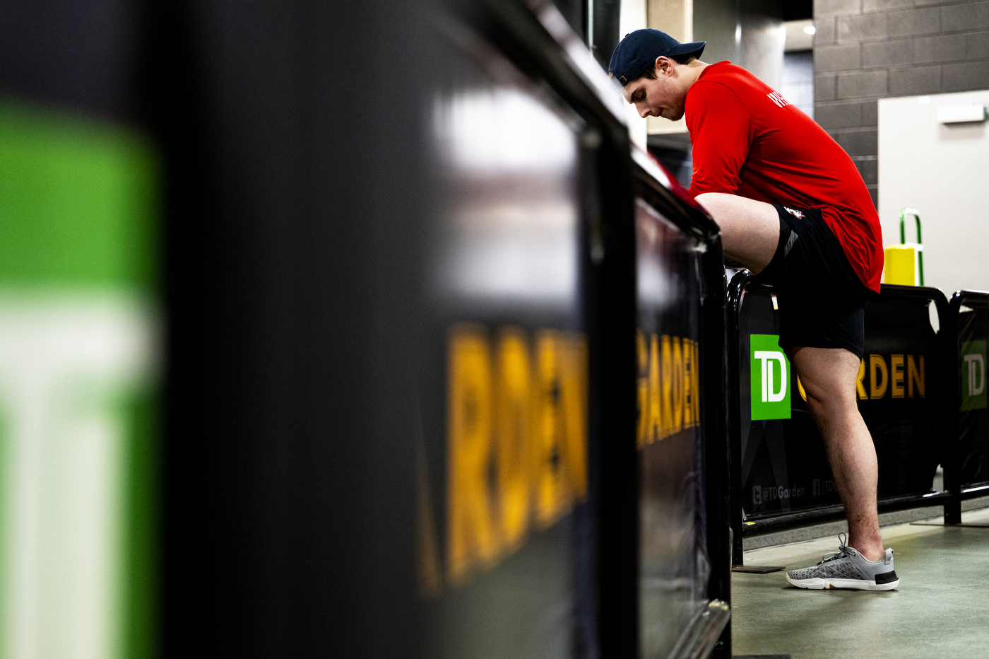 Men's hockey player in red shirt and black shorts and black backwards baseball cap stretching before the game.