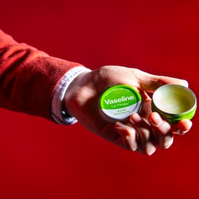 A person holding a tin of Vaseline lip balm in their hand in front of a red background.