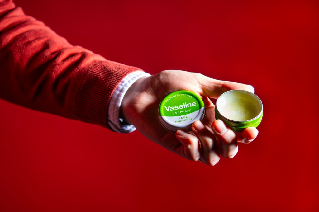 A person holding a tin of Vaseline lip balm in their hand in front of a red background.