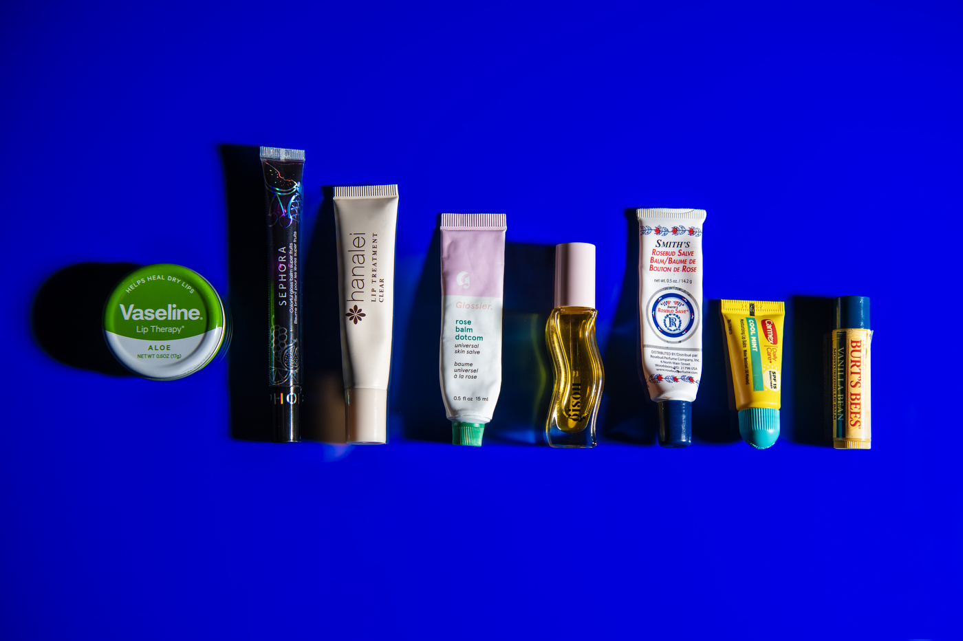 A row of lip products, from left to right: a tin of Vaseline, a tube of Sephora lip balm, a tube of Hanalei lip balm, a tube of Glossier lip balm, a tube of Gisou lip balm, a tube of Smith's lip balm, a tube of Carmex, and a Burt's Bees lip balm.