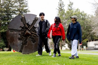 Three people walk past a sculpture outside on a cloudy day.
