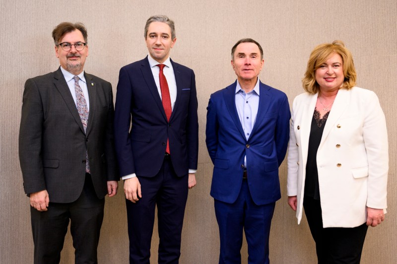 Four people in suits pose for a photo in front of a beige-colored wall.