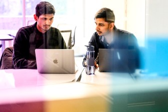 Two people sit in front of laptops in a white-colored room with red, yellow, and green lights.