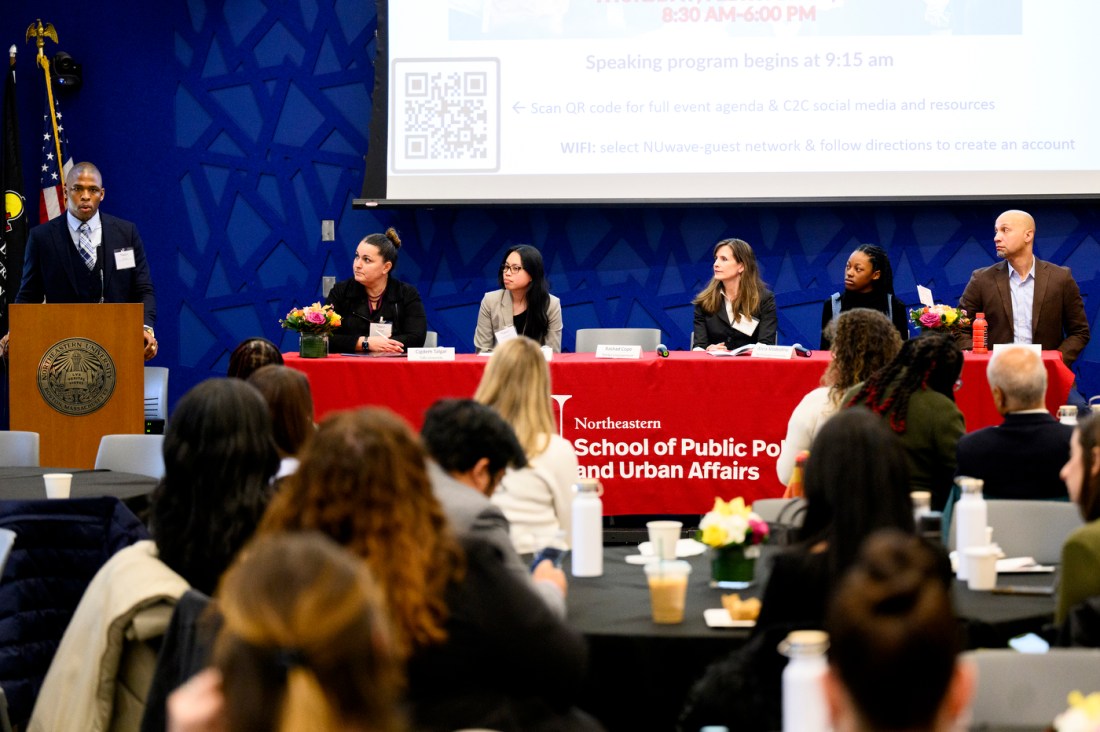 At the table with a red tablecloth, five individuals are seated while a speaker addresses an audience to their left from a podium.