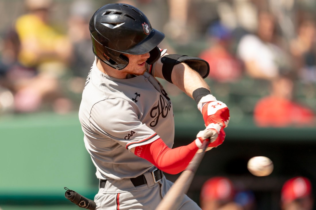 A Northeastern men's baseball player swings a bat to hit a baseball. 