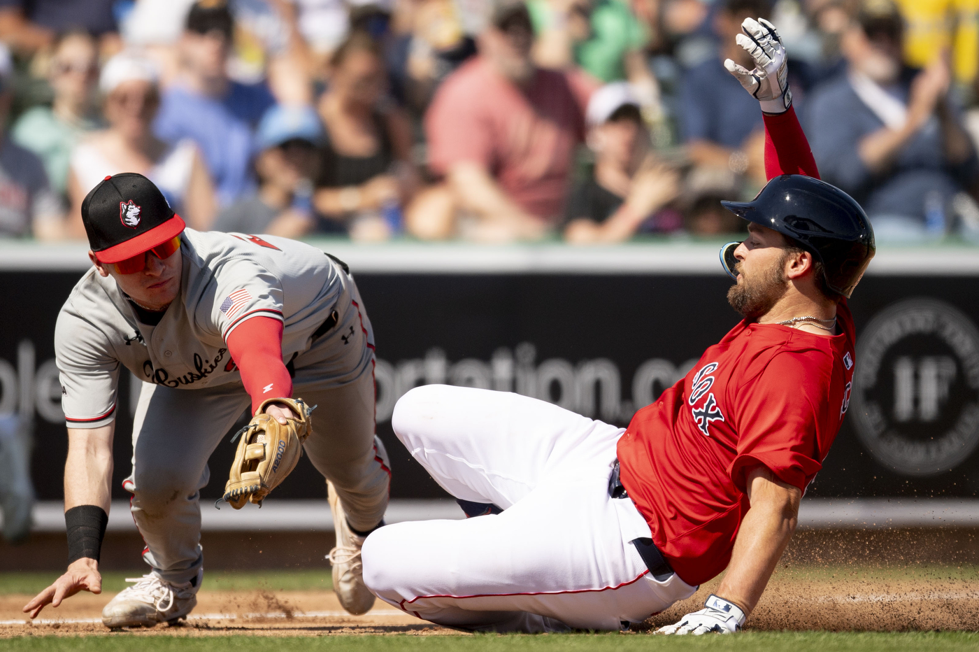 A baseball player wearing a red jersey labeled "Red Sox" slides into a base as a Northeastern baseball player wearing a grey uniform tries to strike them out.