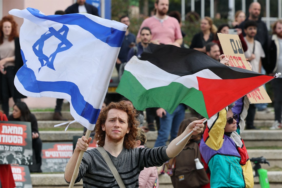 A person in a crowd holds an Israeli flag in one hand an a Palestinian flag in the other hand.