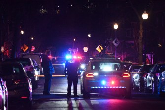 A police officer and another person stand outside in the middle of a street near a police cruiser, flashing red and blue lights.