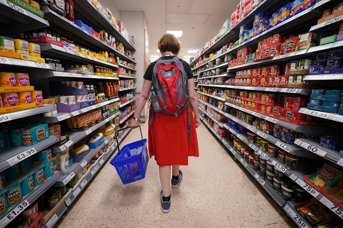 A shopper wearing a backpack carrying a blue basket through the aisles of a supermarket.