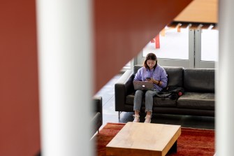 A person wearing a purple sweater works on a laptop while sitting on a black couch.