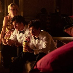 A screen capture of a scene from Saltburn with two men sitting on a couch and a woman standing behind them leaning over them.