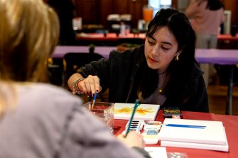 A person sitting in front of another person at a dark orange-colored table paints on a canvas.