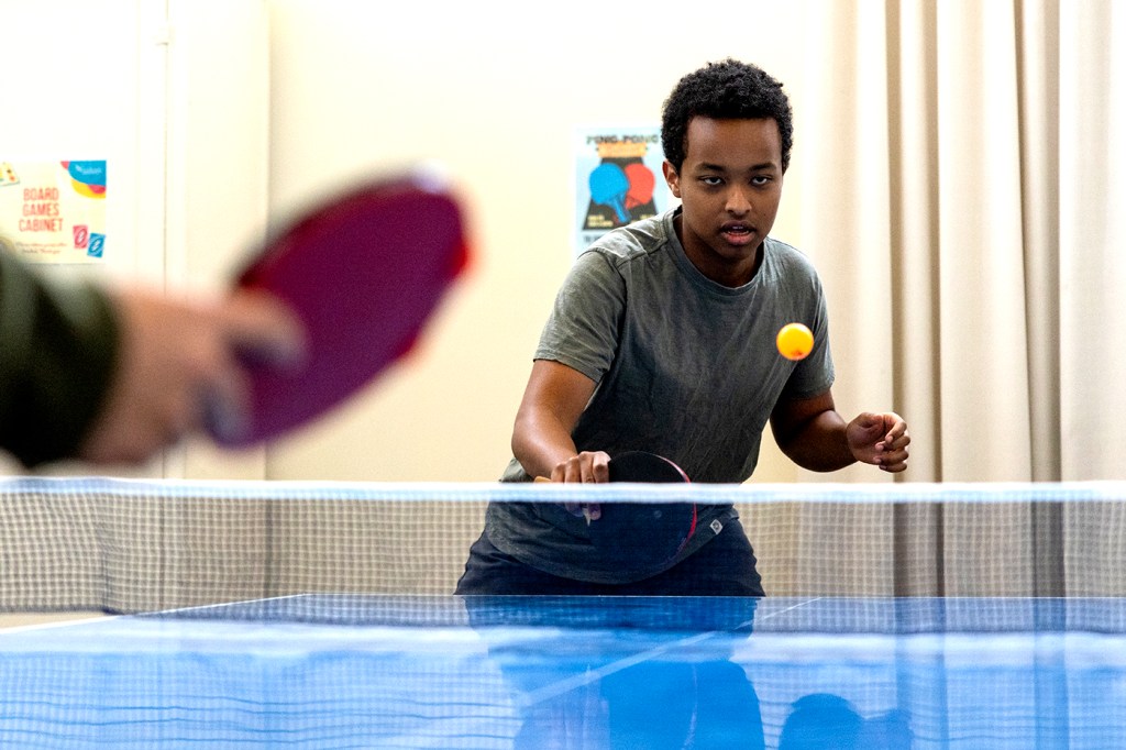 A person wearing a grey shirt and black pants plays table tennis against an opponent.