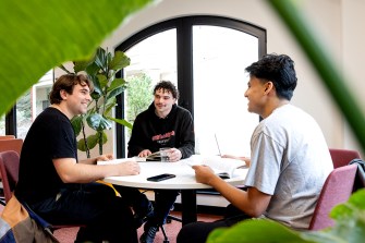 Three people sit around a table, working on a project in a brightly lit, white-colored room.