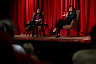 Two people talk to an audience while sitting in separate chairs on a stage in front of large red-colored curtains.