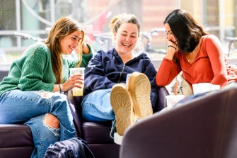 Three students sit on chairs as they look at one person's phone smiling and laughing.