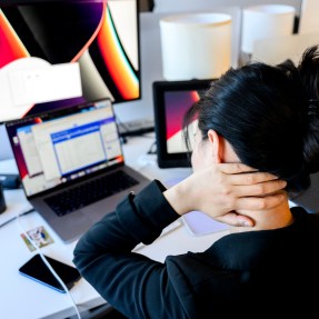 A person sitting at a desk in front of a laptop and dual monitors holding the back of their neck.