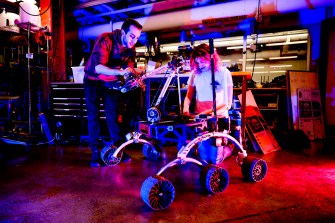 Two students, one standing and one kneeling, work on a robotic rover in the Richards Hall Makerspace. They are bathed in purple and red light.