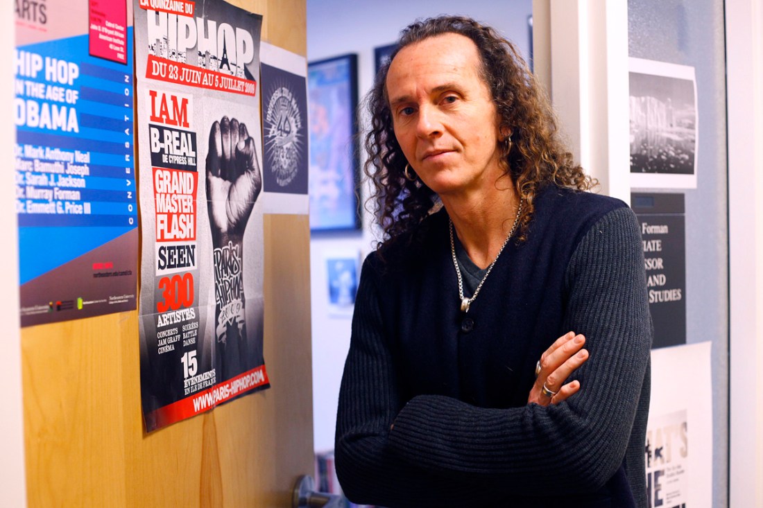 A man with long hair and dark clothes poses next to an office with hip-hop fliers affixed to the door.